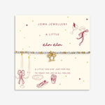 A beaded bracelet with a gold bow charm displayed on a cream-colored Joma Jewellery card. The bracelet features white and gold beads with a central gold bow charm.