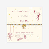 A beaded bracelet with a gold bow charm displayed on a cream-colored Joma Jewellery card. The bracelet features white and gold beads with a central gold bow charm.