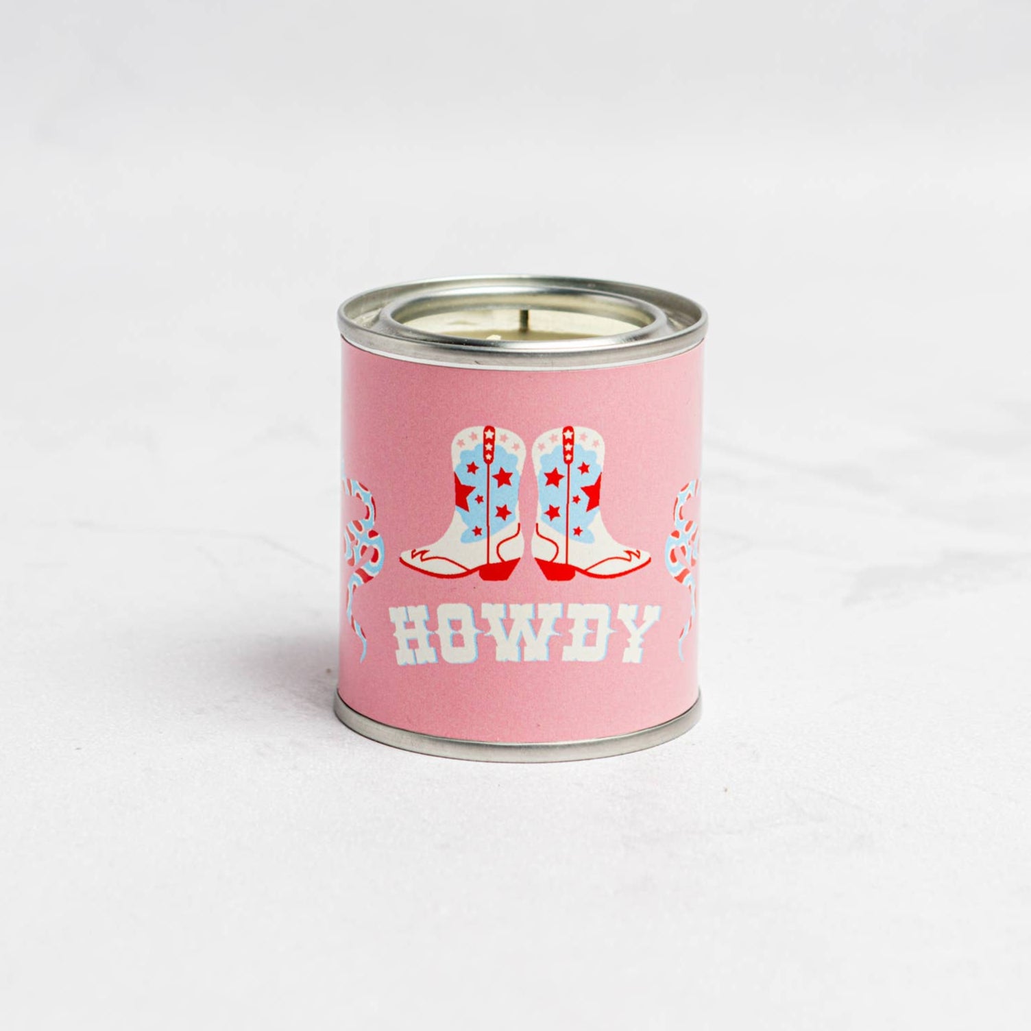 A pink candle with 'Howdy' text and decorative elements in a cowboy hat and boots on the label, placed on a white background.
