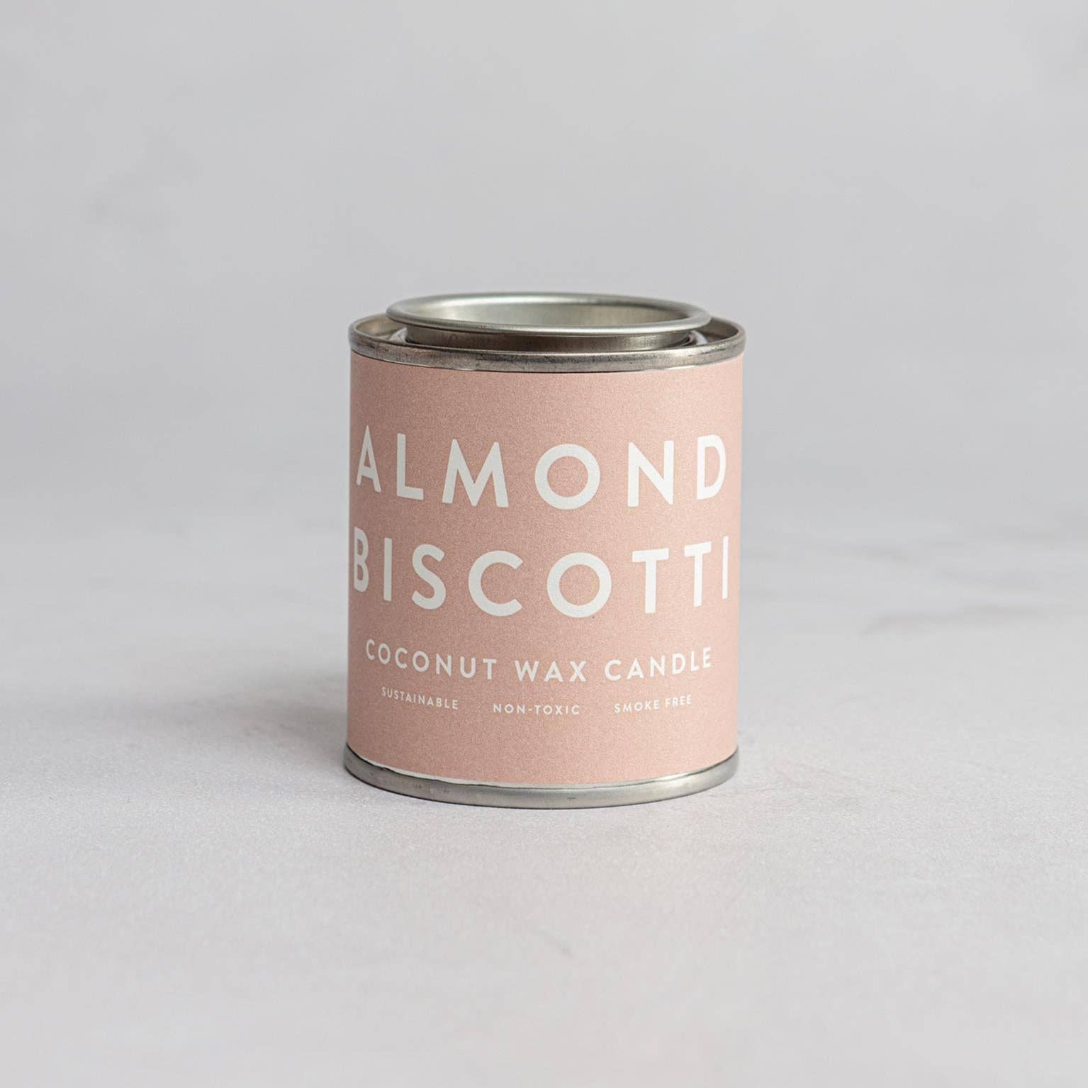A cylindrical pink-labeled Almond Biscotti coconut wax candle in a recyclable metal tin.