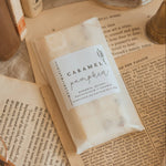 A packaged food product labeled 'Caramel Pumpkin' resting on open vintage-style books with a brass candle holder and fabric in the background.