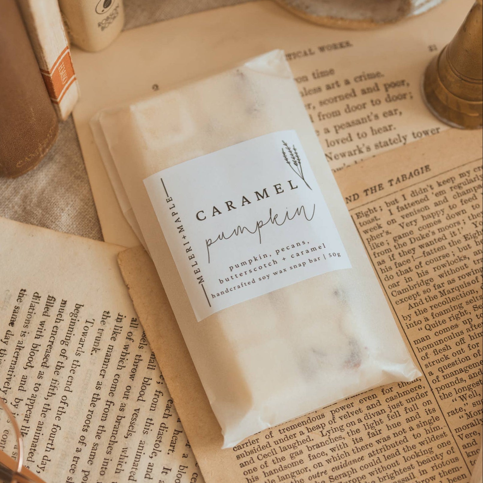 A packaged food product labeled 'Caramel Pumpkin' resting on open vintage-style books with a brass candle holder and fabric in the background.