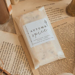 A clear plastic bag labeled 'Autumn Spice' filled with pale beige spice blends, styled on top of open vintage-style books with a small brass candle holder and cream fabric in the background.