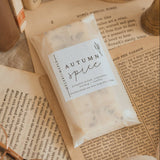 A clear plastic bag labeled 'Autumn Spice' filled with pale beige spice blends, styled on top of open vintage-style books with a small brass candle holder and cream fabric in the background.