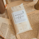 A small white bag labeled 'MORNING coffee' with 'fresh coffee beans + coconut' and 'handcrafted one cup at a time' lies on top of open vintage-style books and paper, with a textured ceramic bowl and soft fabric around it.