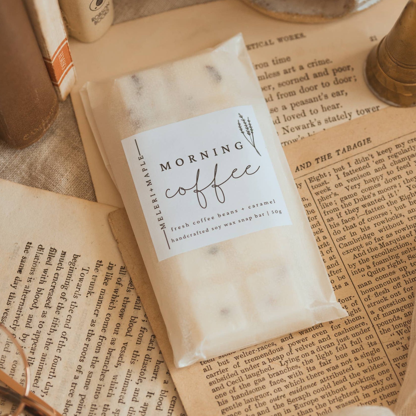 A small white bag labeled 'MORNING coffee' with 'fresh coffee beans + coconut' and 'handcrafted one cup at a time' lies on top of open vintage-style books and paper, with a textured ceramic bowl and soft fabric around it.