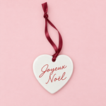 A white ceramic Christmas ornament shaped like a heart, featuring the French greeting 'Joyeux Noel' in red cursive script, hanging from a dark red ribbon against a solid light pink background.