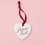 A white ceramic Christmas ornament shaped like a heart, featuring the French greeting 'Joyeux Noel' in red cursive script, hanging from a dark red ribbon against a solid light pink background.