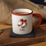 A white ceramic mug with a reddish-brown handle and rim, decorated with two orange-and-white knit socks and the words 'Snuggle Season,' set on a dark coaster over a rustic wooden surface with autumn leaves, burlap, and small pumpkins around it.
