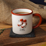 A white ceramic mug with a reddish-brown handle and rim, decorated with two orange-and-white knit socks and the words 'Snuggle Season,' set on a dark coaster over a rustic wooden surface with autumn leaves, burlap, and small pumpkins around it.