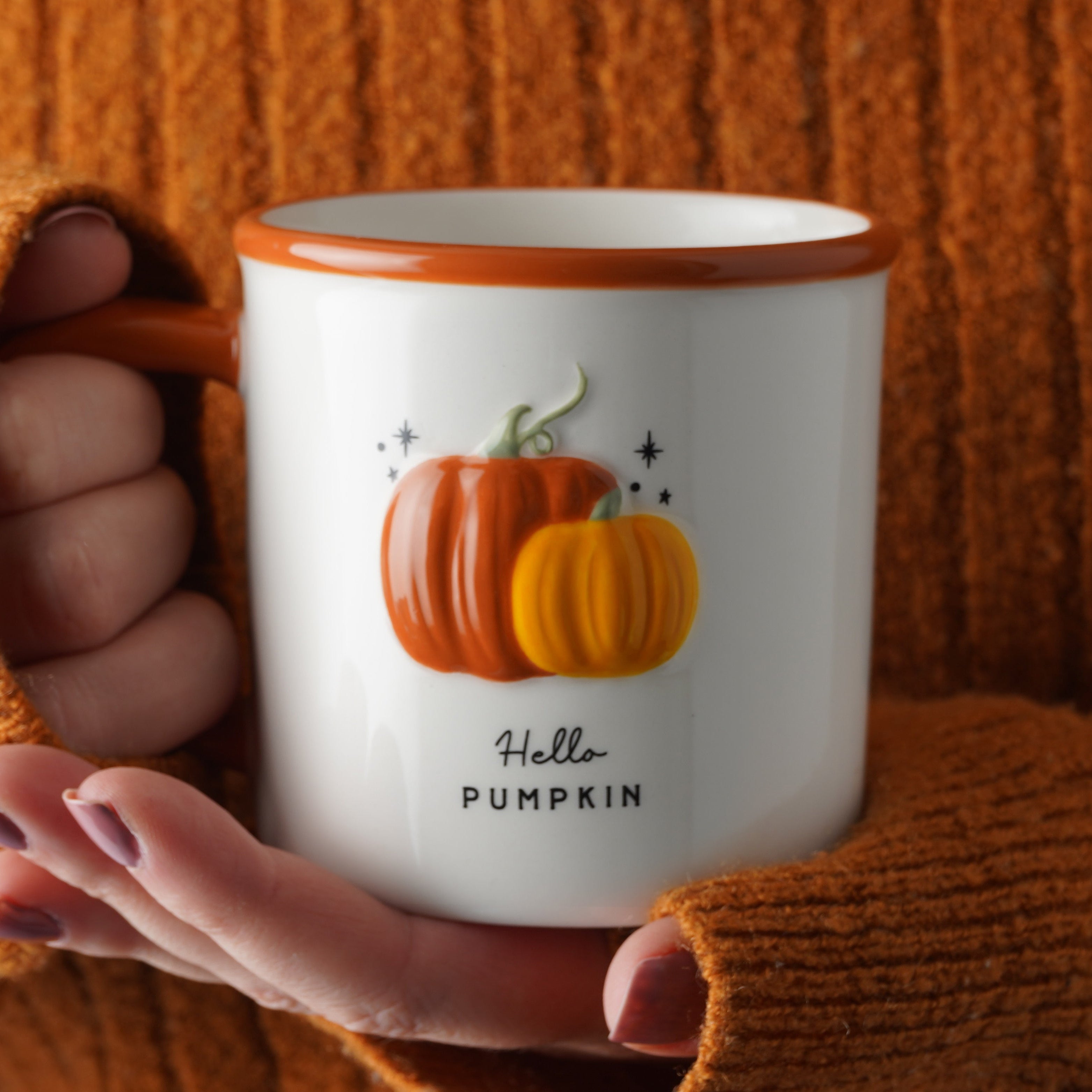 A white ceramic mug with a brown rim and handle, featuring a graphic of two pumpkins and the text 'Hello PUMPKIN', held by hands wearing an orange ribbed knit sweater against a matching orange background.
