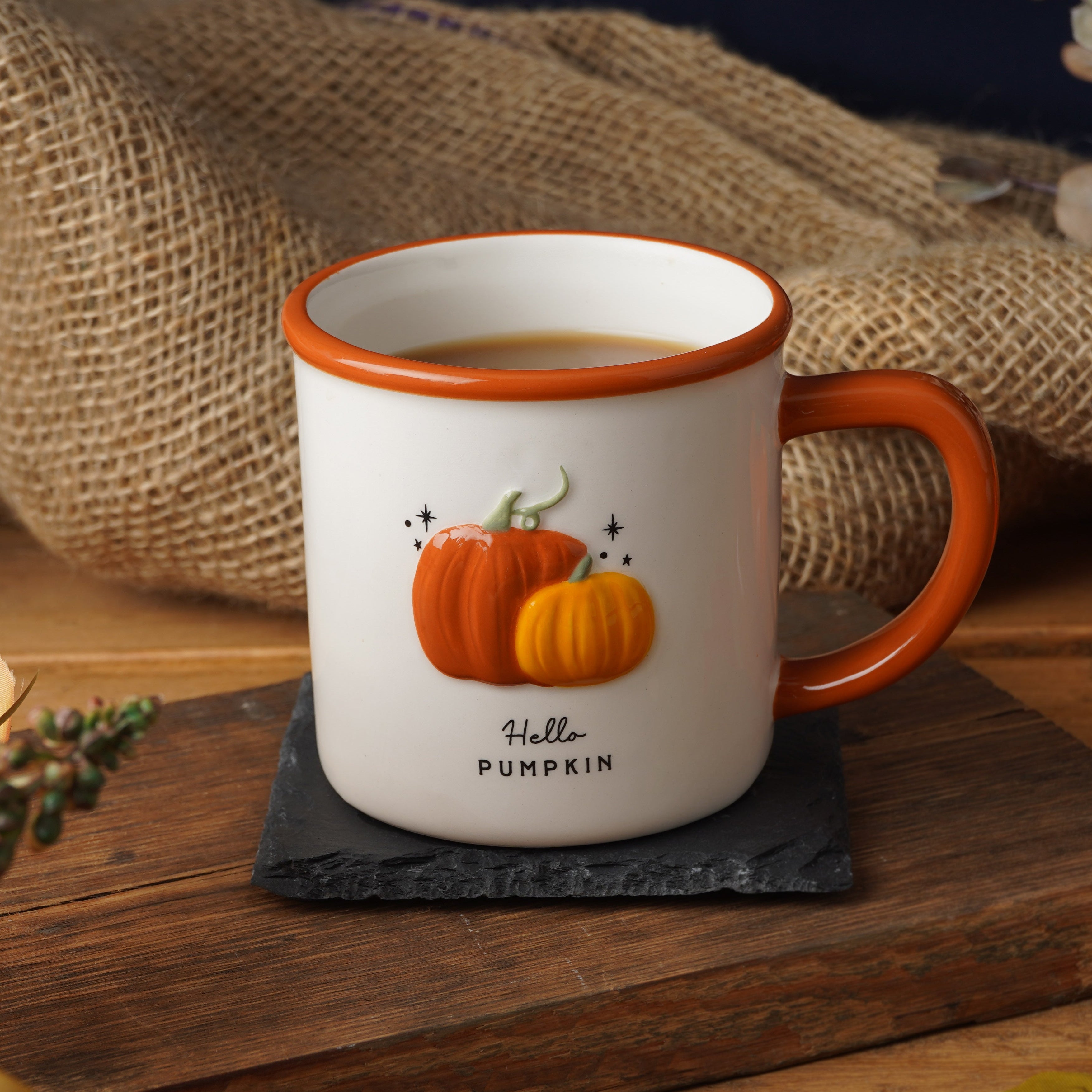 A white ceramic coffee mug with an orange handle and rim, decorated with two orange pumpkins and the text 'Hello PUMPKIN', sitting on a dark slate coaster on a rustic wooden table surrounded by autumn leaves, burlap, and small pumpkins.