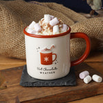 A white ceramic mug with a red handle and rim, decorated with a red mug graphic and the words 'Hot Chocolate Weather'. The mug is filled with whipped cream, marshmallows, and a candy cane, set on a dark slate coaster on a wooden table with burlap and dried flowers in the background.