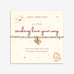 A Joma Jewellery bracelet on a cream gift card. The bracelet features a gold charm with a red heart and a silver and white beaded band.