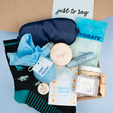 A curated gift box flat-lay featuring a navy blue sleep mask, black socks with teal and gray animal prints, a light blue scrunchie, a round cream makeup sponge with 'kugls' written on it, a tube of before sleep serum, a packet of blue agave hydrating mask, a packet of eggstra special exfoliating scrub, and a jar of bath crystals, arranged on a light blue background with a kraft box and a card that reads 'just to say'.