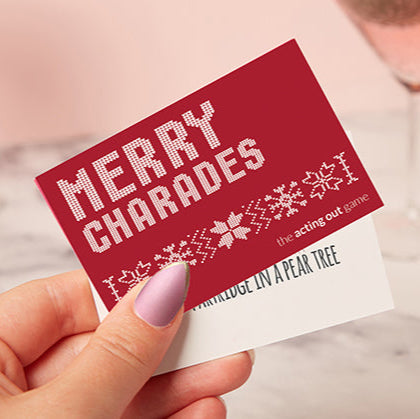 Hand holding a red card with 'Merry Charades' text and decorative elements.