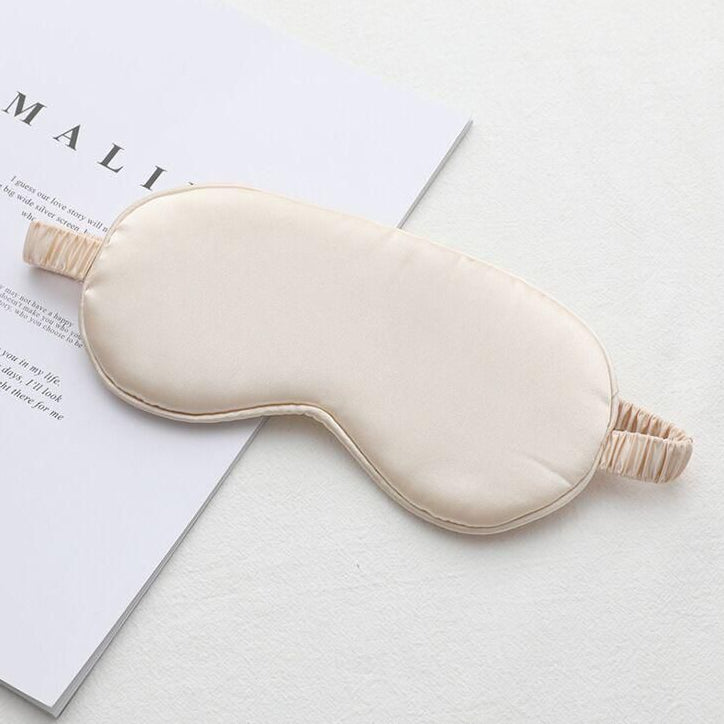 A cream satin eye mask on a pale background, with a book or paper in the background.