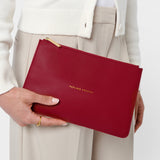 Red clutch bag held by a person wearing a white sweater and beige pants on a light gray background